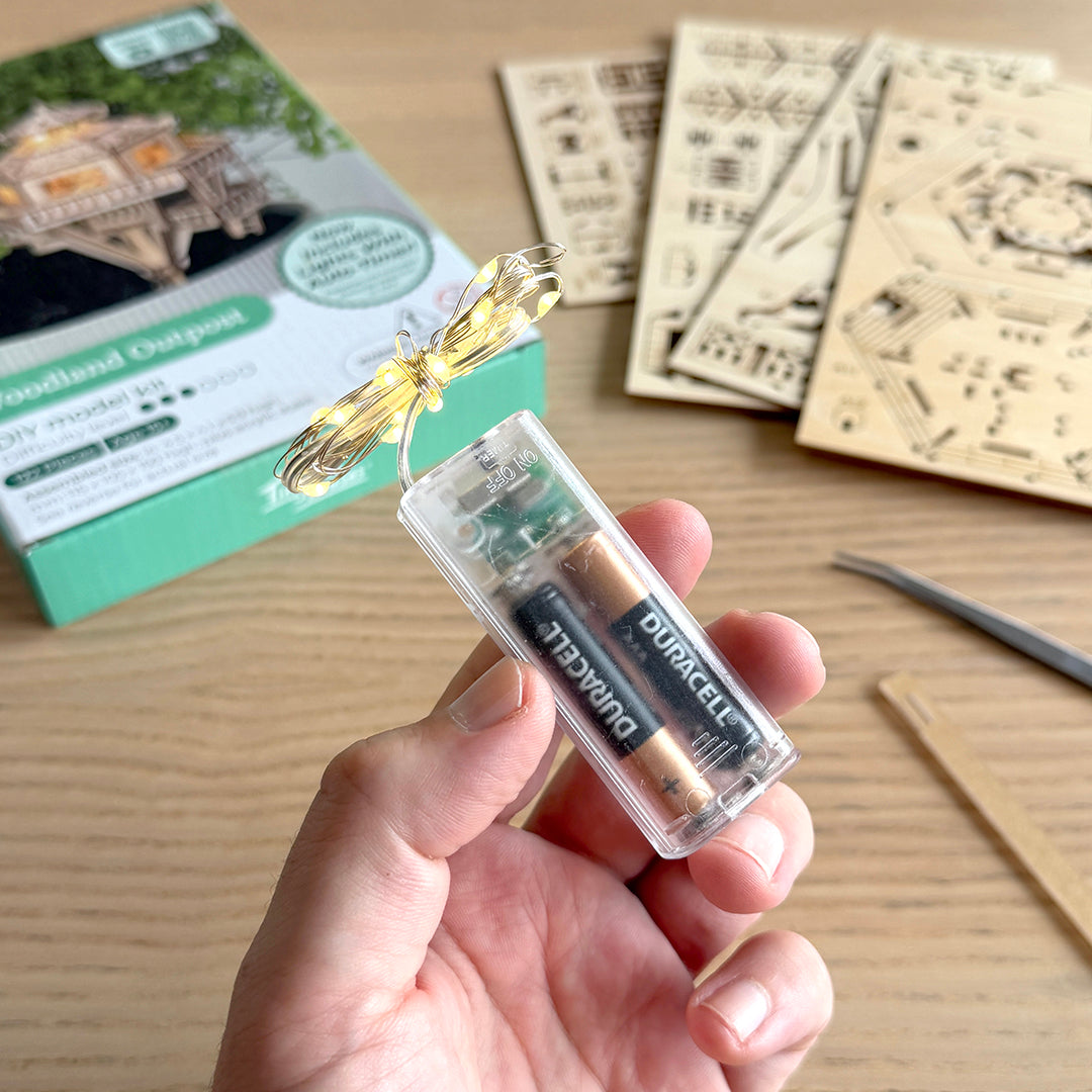 A hand holds a small clear plastic battery-operated light string with warm yellow LEDs and a visible Duracell battery inside. In the background on a wooden table is the product box, laser-cut wooden model pieces, tweezers, and other kit components.