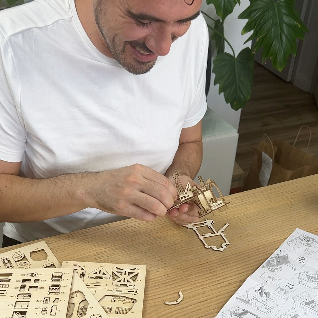Lars Wijers, creator of Tiny Treehouses  wearing a white t-shirt is shown assembling a wooden laser-cut Tiny Treehouse model on a light wood table, with parts and an instruction sheet visible.