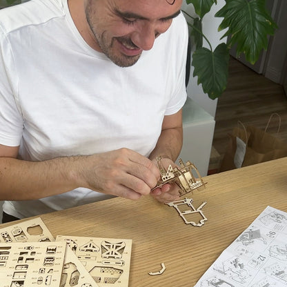 Lars Wijers, creator of Tiny Treehouses  wearing a white t-shirt is shown assembling a wooden laser-cut Tiny Treehouse model on a light wood table, with parts and an instruction sheet visible.