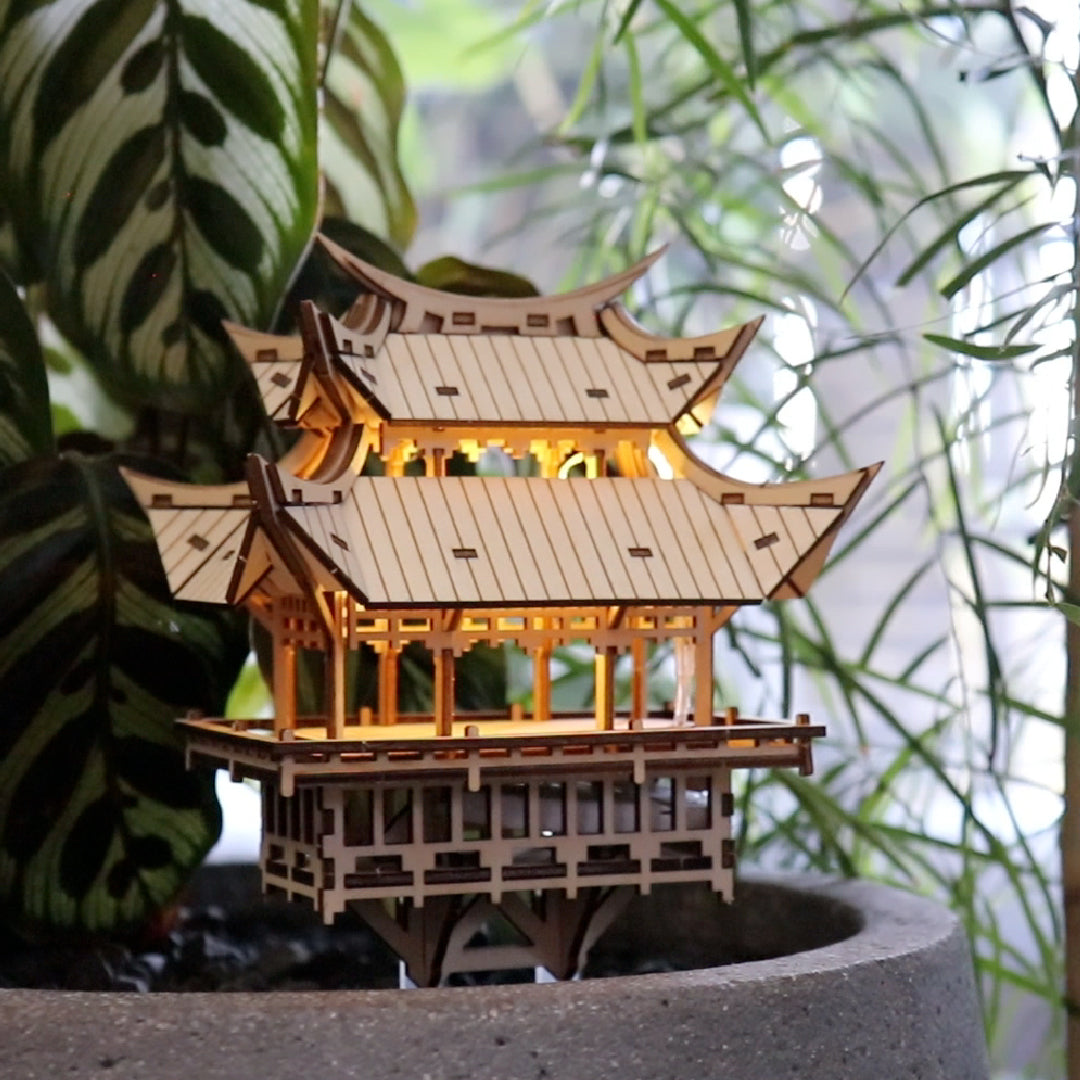 Tiny Treehouses 3D Puzzle Wooden Tiny Treehouse 3D Puzzles pagoda model with glowing interior lights, placed in a pot with green plants in the background.