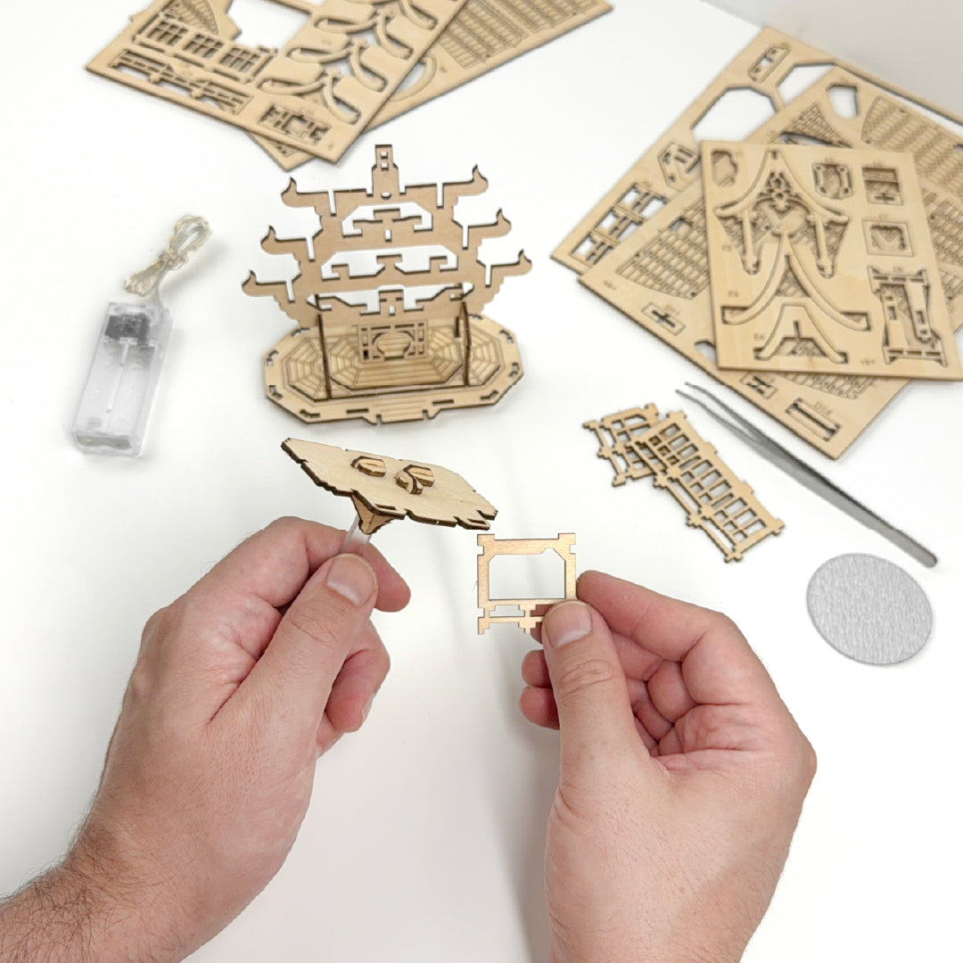 Tiny Treehouses 3D Puzzle Wooden architectural models and tools on a white surface with hands holding a small model.