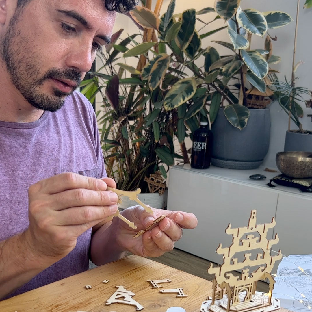 A man in a purple shirt is assembling a Tiny Treehouse 3D puzzle wooden geometric model on a table, with a completed multi-tiered pagoda structure and small wooden pieces nearby. The background features a white cabinet, a black bottle labeled &